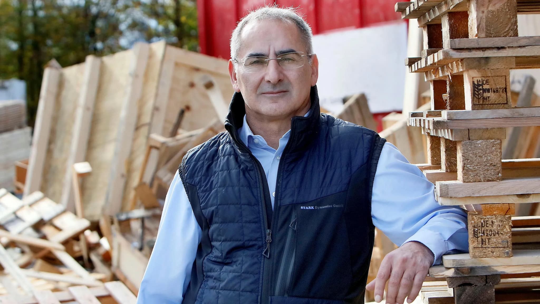 Eine Konzeptzeichnung eines Industrielagers, umgeben von Holzpaletten und Kisten, die mit Baumaterialien gefüllt sind. Der Entwurf enthält detaillierte Illustrationen, die die Anordnung verschiedener Arten von Holzbrettern in unterschiedlichen Größen zeigen, sowie Entwürfe für Türen oder Fenster auf einer Seite des Gebäudes. Außerdem gibt es eine Skizze, die zeigt, wie diese Gegenstände im Inneren des Gebäudes angeordnet werden würden, alles vor einem weißen Hintergrund.